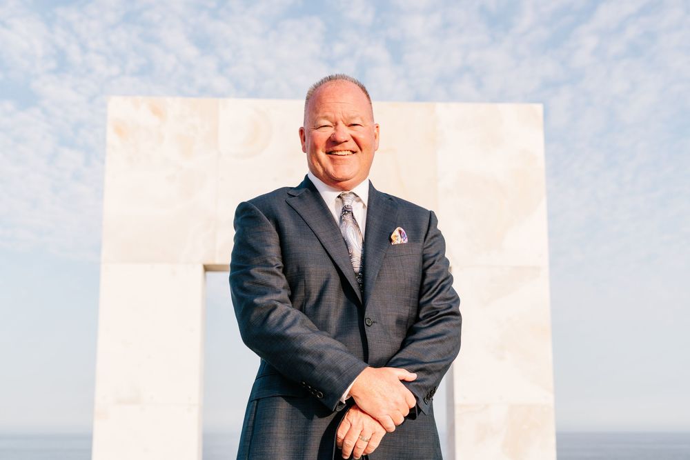 Professional man in navy suit smiling confidently outdoors near white marble monument against blue sky.