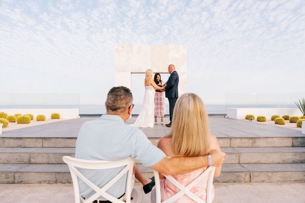 Couple watching an outdoor wedding ceremony at a rooftop venue with ocean views under a bright sky.