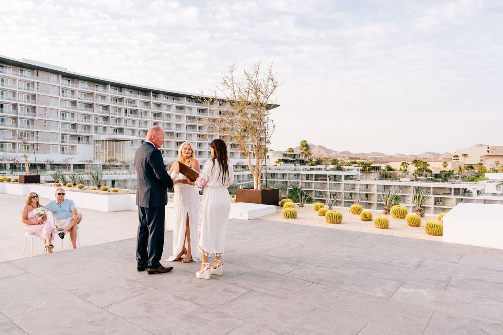 Outdoor rooftop wedding ceremony with couple exchanging vows, officiant, and guests near a modern hotel.