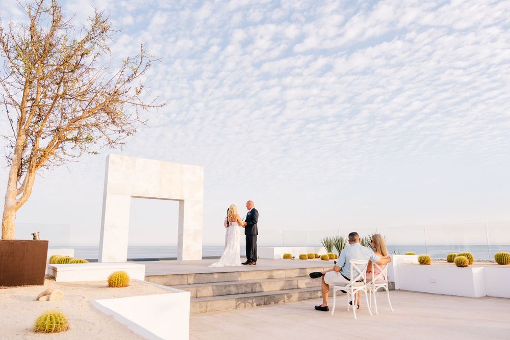 Outdoor wedding ceremony on a rooftop terrace with ocean views, white arch, cactus decor, and blue sky backdrop.