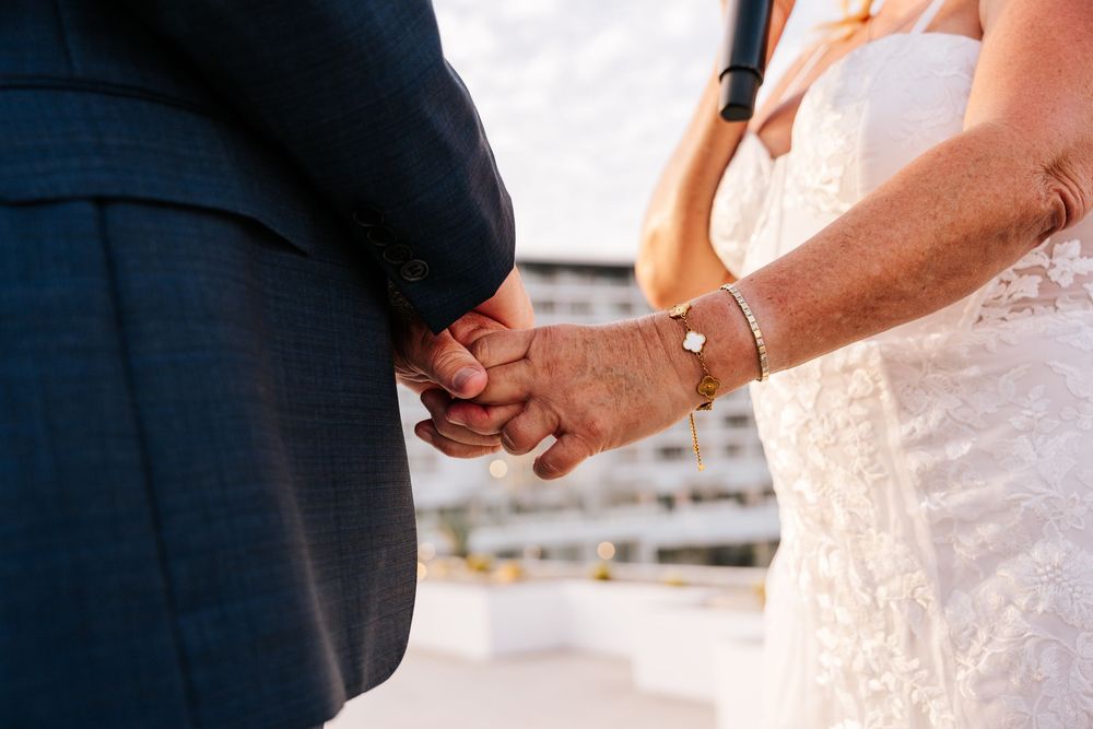 Elderly couple holding hands at wedding ceremony, bride in lace dress with gold bracelet, groom in navy suit.