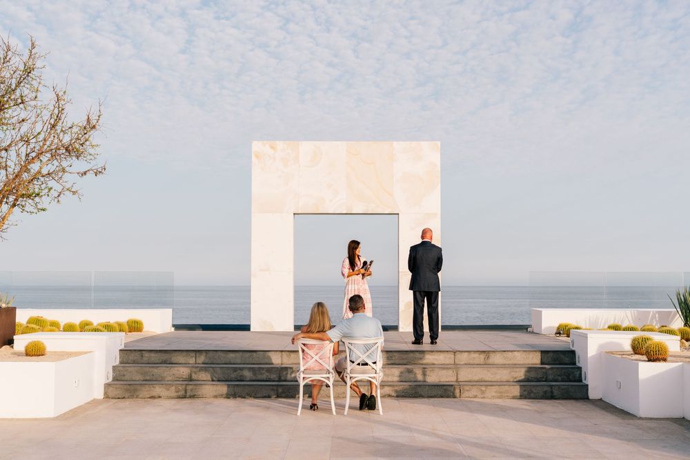Small intimate wedding ceremony on ocean-view terrace with white architectural arch and two seated guests.