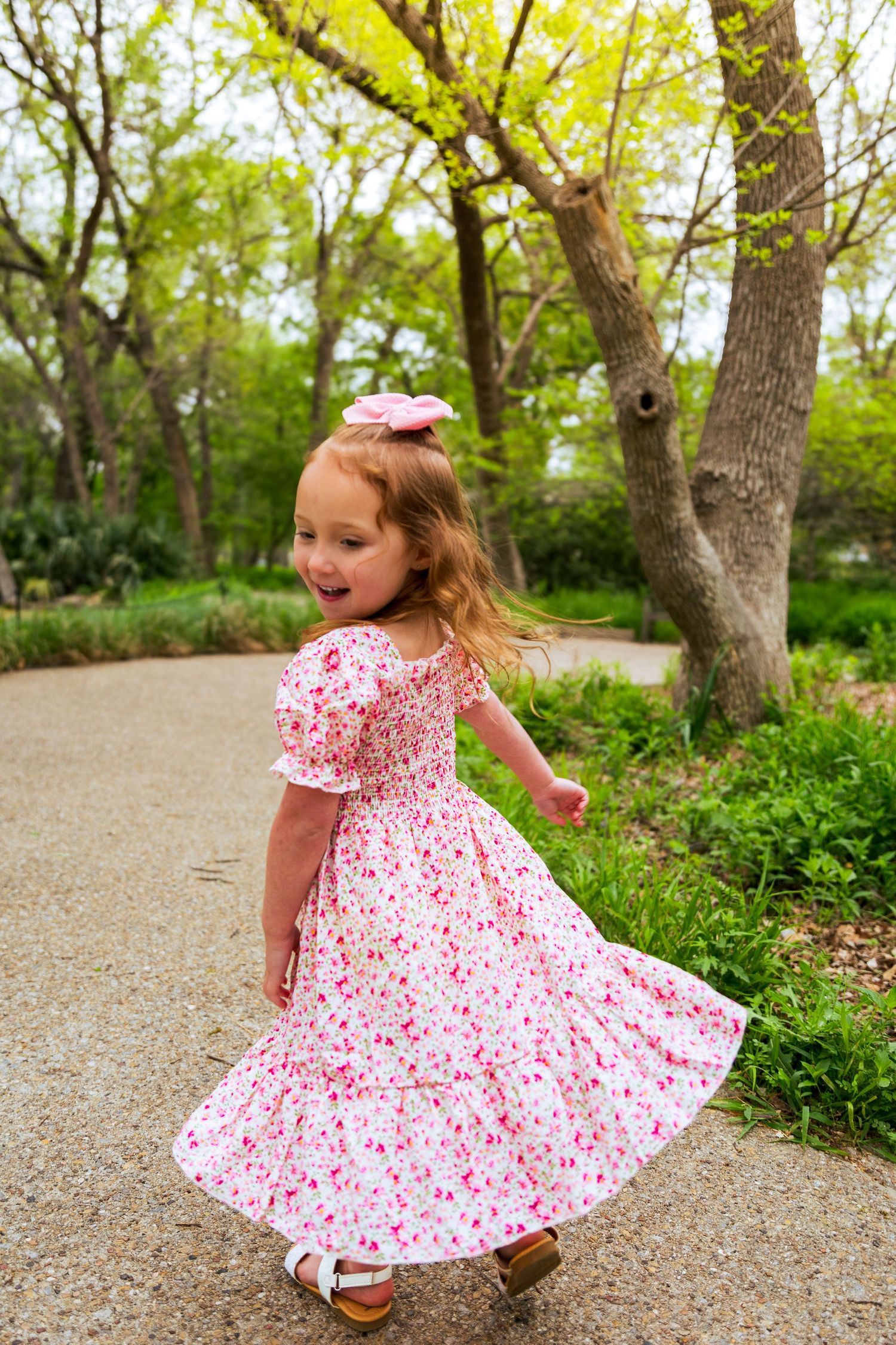 Young girl twirling in a pink floral dress with a bow in her hair on a park path surrounded by green trees.