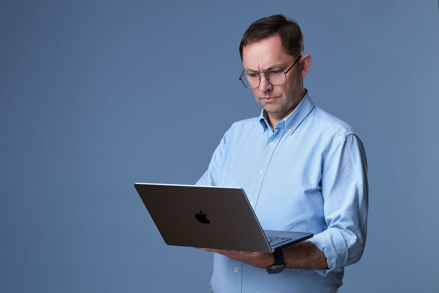 Middle-aged man in blue shirt and glasses holding an Apple MacBook laptop against a blue background.