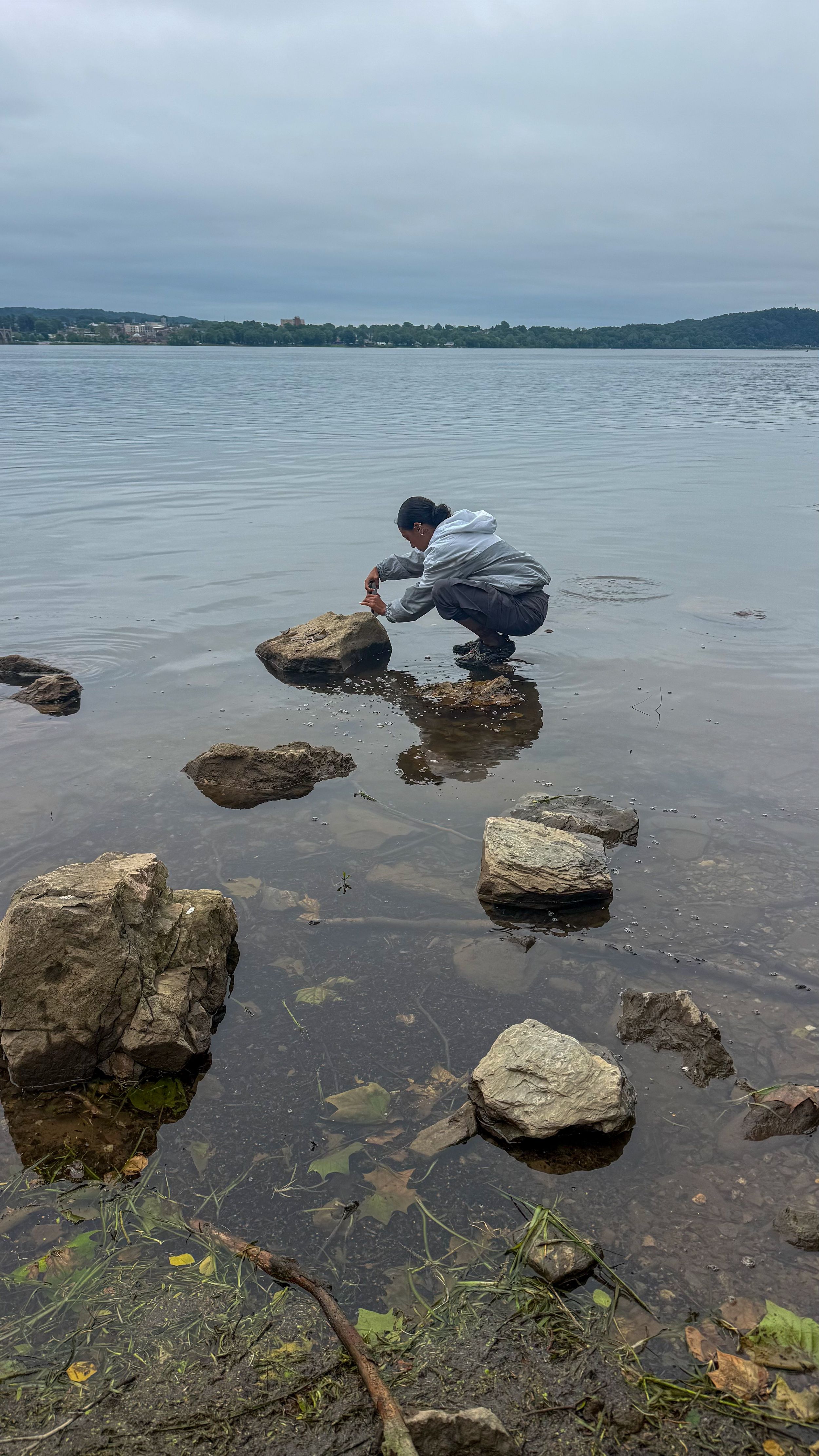 Person crouching on rocks at water's edge examining stones in a calm lake under overcast skies.