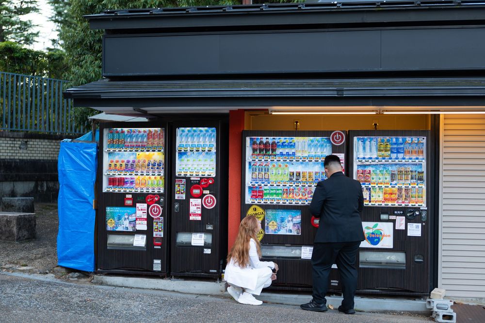 Couple standing before a row of Japanese vending machines outdoors during daytime.