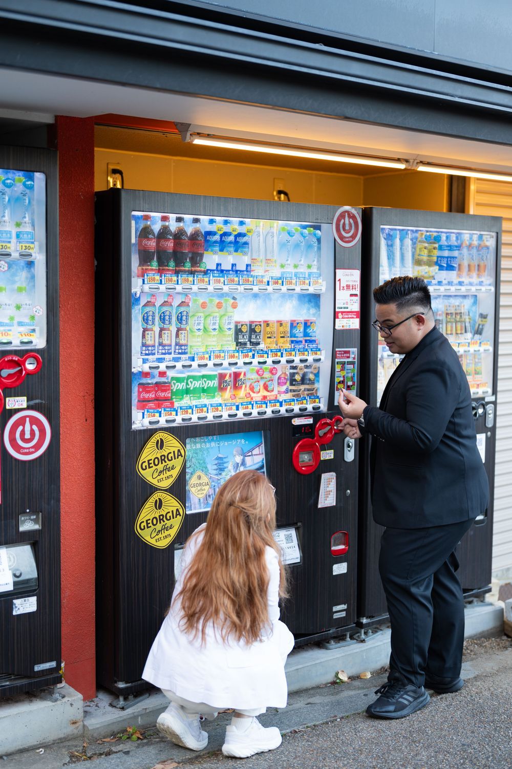 Man in black suit and woman in white crouching at a Japanese vending machine street corner.