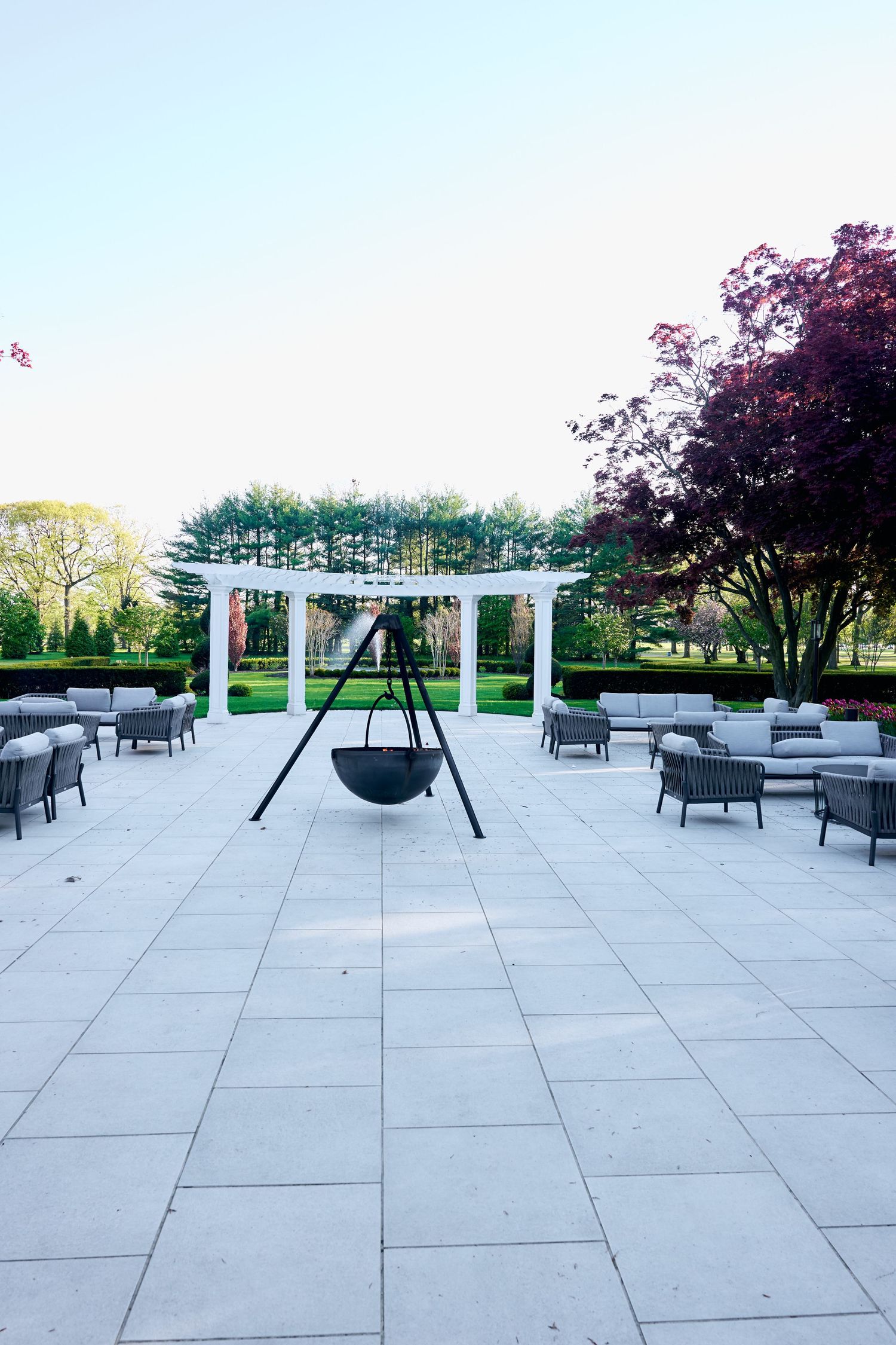 Elegant outdoor patio with gray lounge furniture, tripod fire bowl, white pergola, and Japanese maple tree in manicured garden.