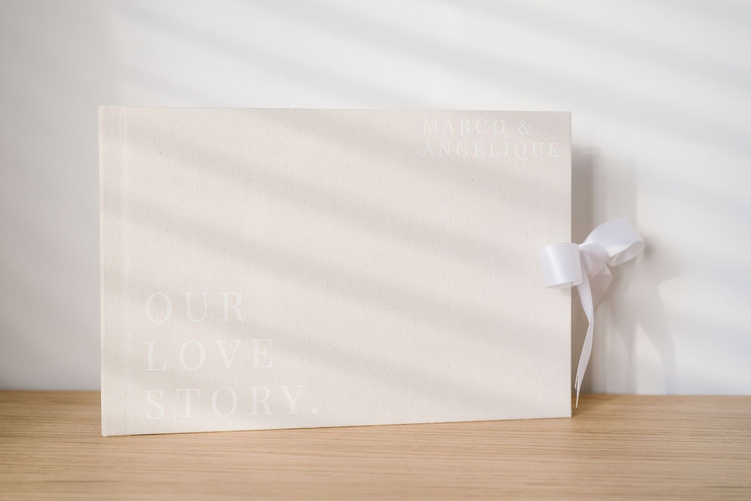 Elegant white wedding photo album with embossed Our Love Story text and satin ribbon bow on wooden surface.
