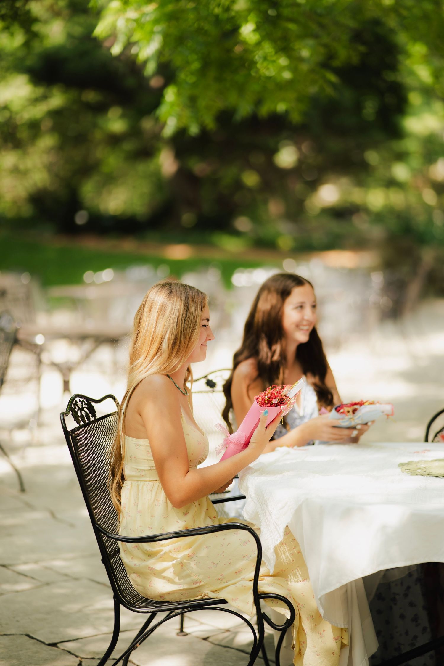 Two young women in summer dresses smiling while seated at an outdoor garden party table with gifts.