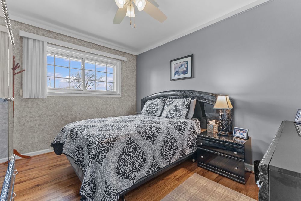Cozy bedroom with black upholstered bed, patterned bedding, hardwood floors, ceiling fan, and neutral gray walls with wallpaper accent.