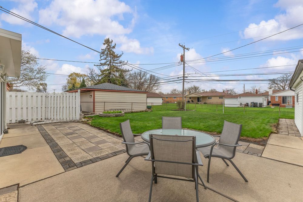 Spacious backyard patio with glass table and chairs, lush green lawn, wooden fence, and utility shed under a blue sky.