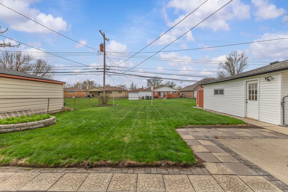 Spacious backyard with lush green lawn, patio pavers, shed, and privacy fence under a partly cloudy sky.