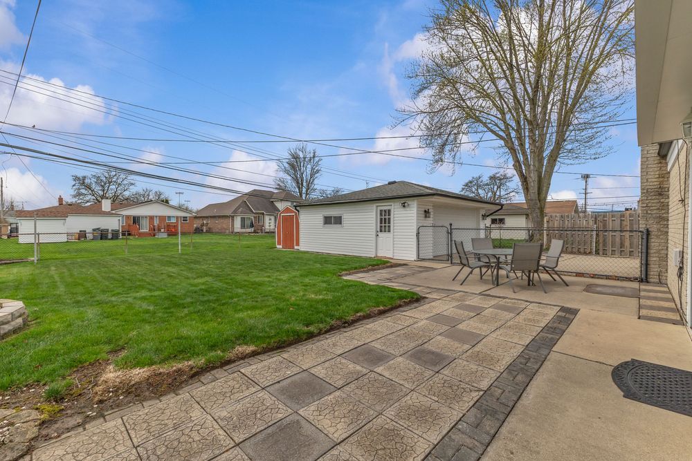 Spacious backyard with lush green lawn, patio dining set, detached garage, and large tree under a bright blue sky.