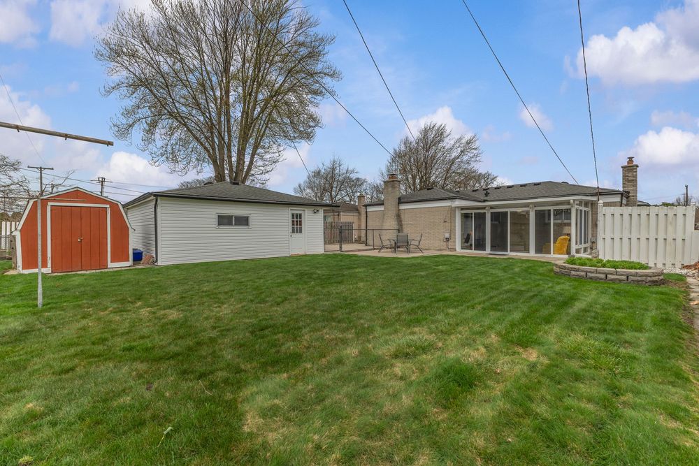 Spacious backyard with lush green lawn, red storage shed, sunroom addition, and white privacy fence on a cloudy day.