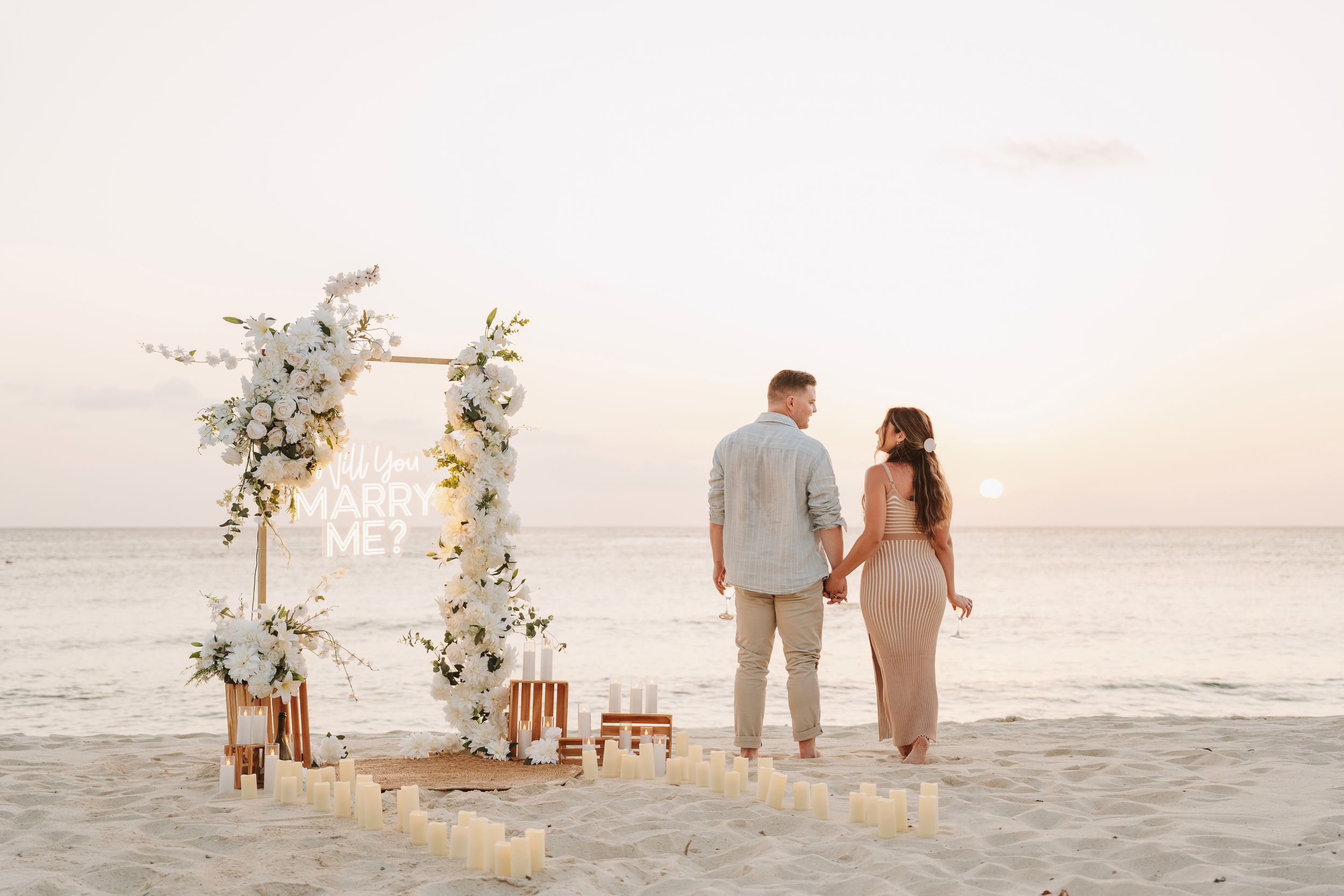 Couple holding hands at sunset beach proposal setup with floral arch, candles, and Marry Me sign on sandy shore.