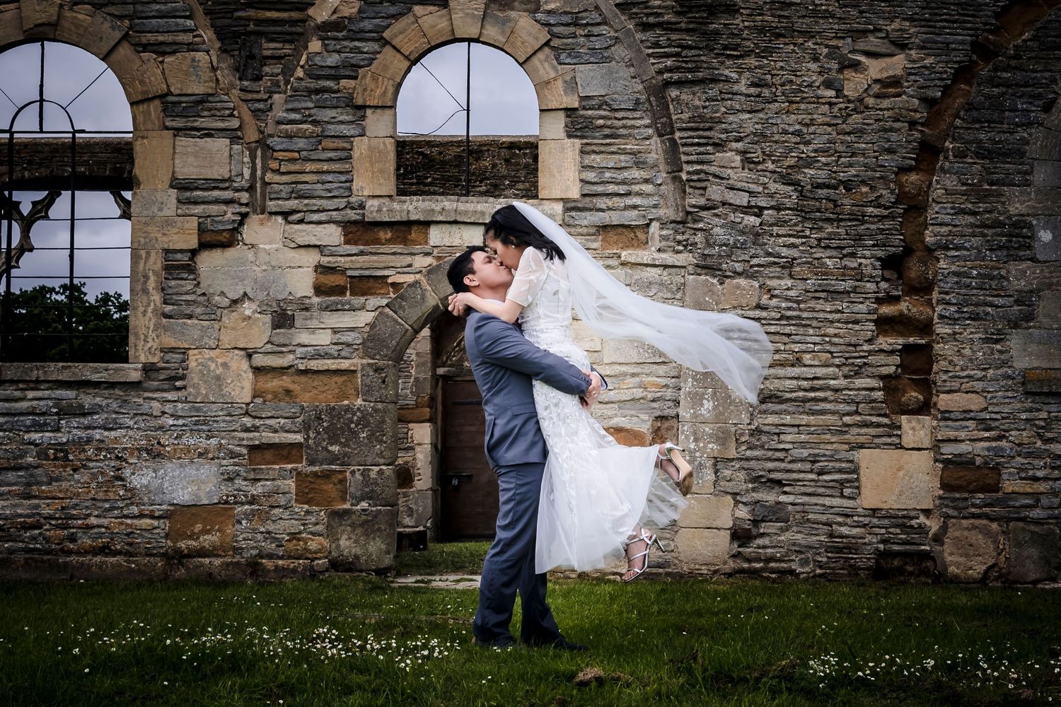 Groom lifts bride in wedding attire outside a rustic stone church, her veil flowing in the breeze.