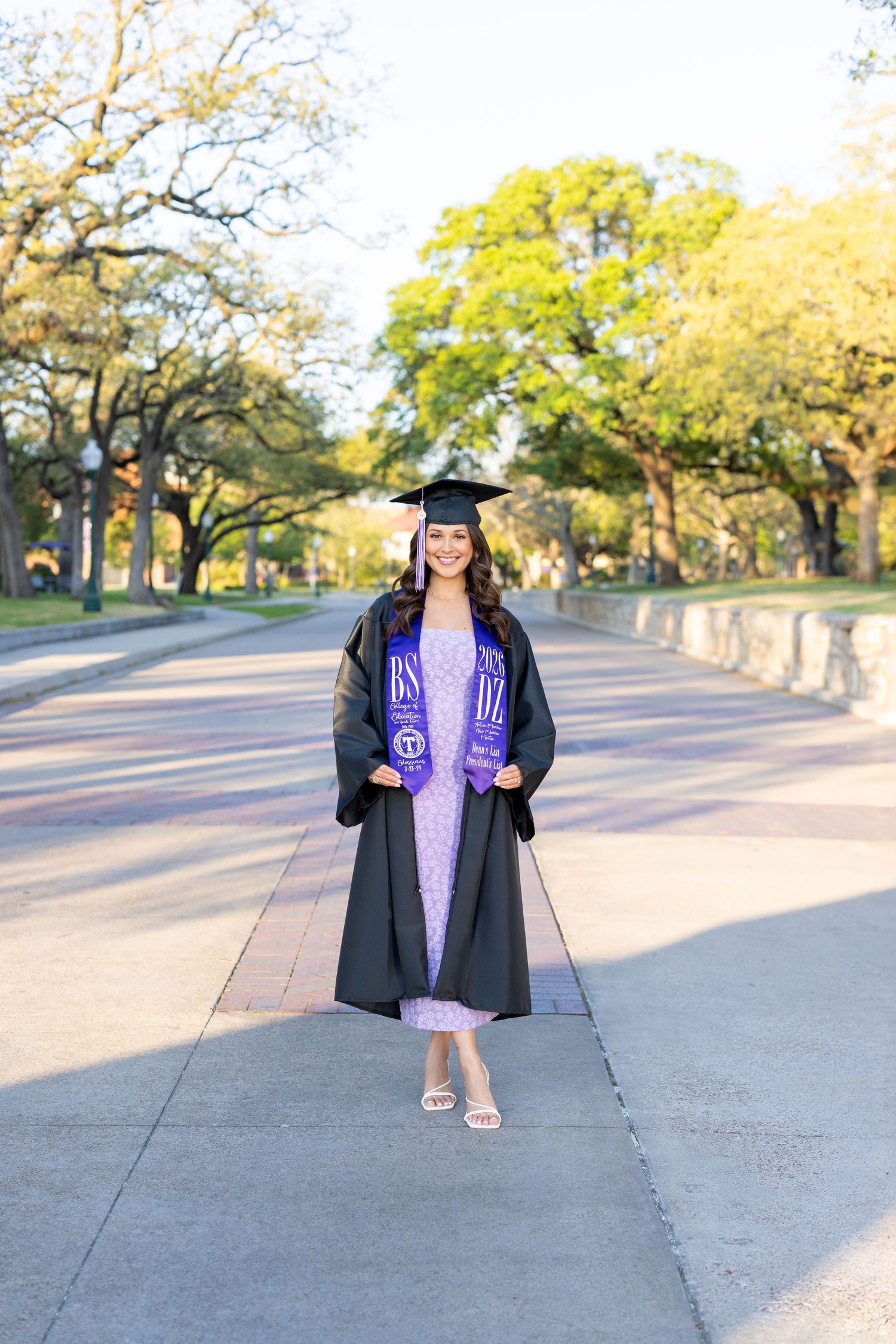 A smiling graduate in black cap and gown with purple stole poses on a tree-lined campus walkway.