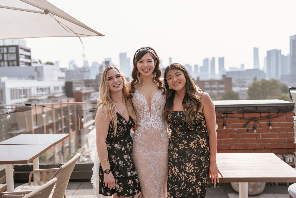 Bride in lace gown poses with two guests on a rooftop venue with city skyline views.