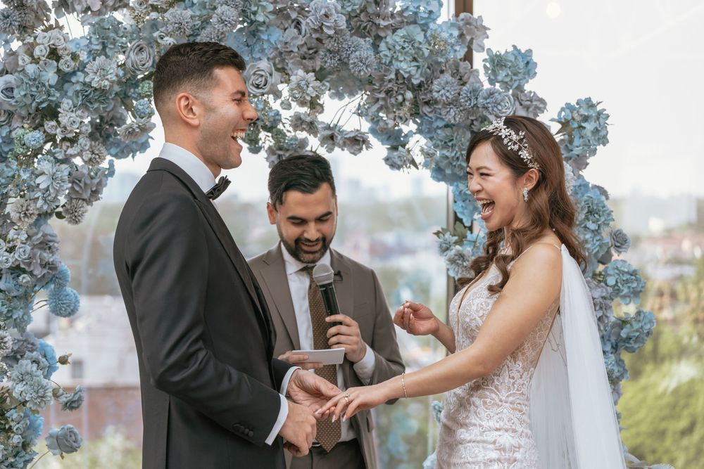 Joyful wedding ceremony with couple exchanging rings under blue hydrangea arch, officiant smiling beside them.