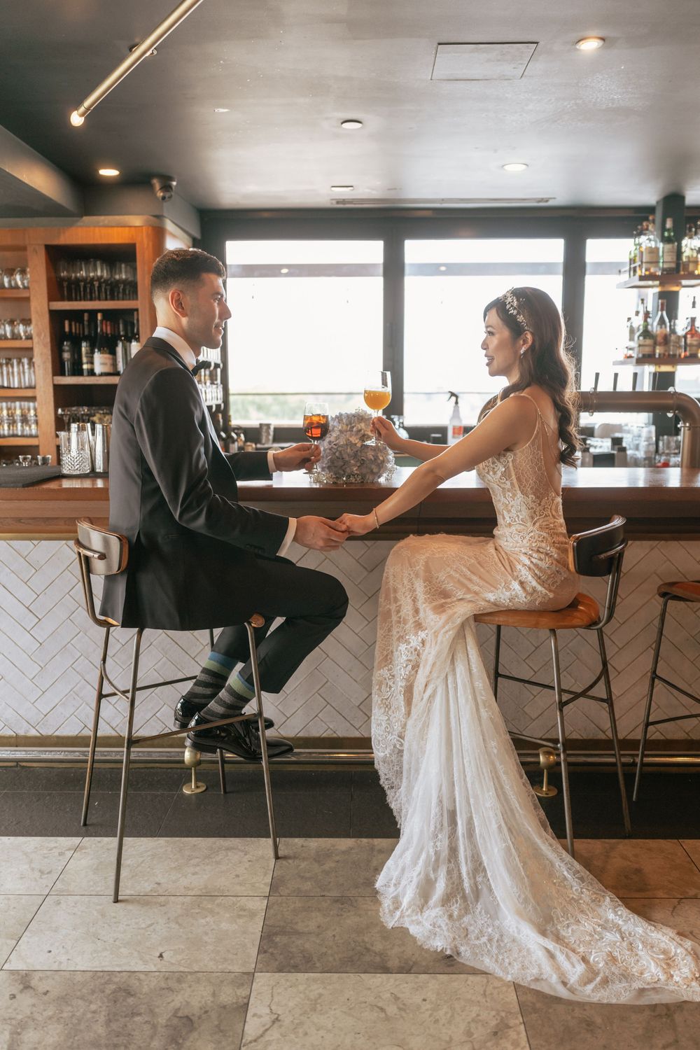Bride in lace gown and groom in black suit sit at a bar holding hands, sharing a romantic wedding day moment.