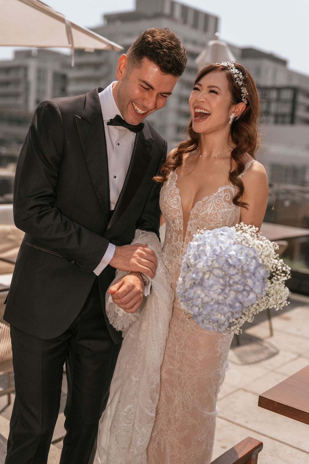 Laughing bride and groom in black tuxedo and lace wedding dress holding hydrangea bouquet outdoors.