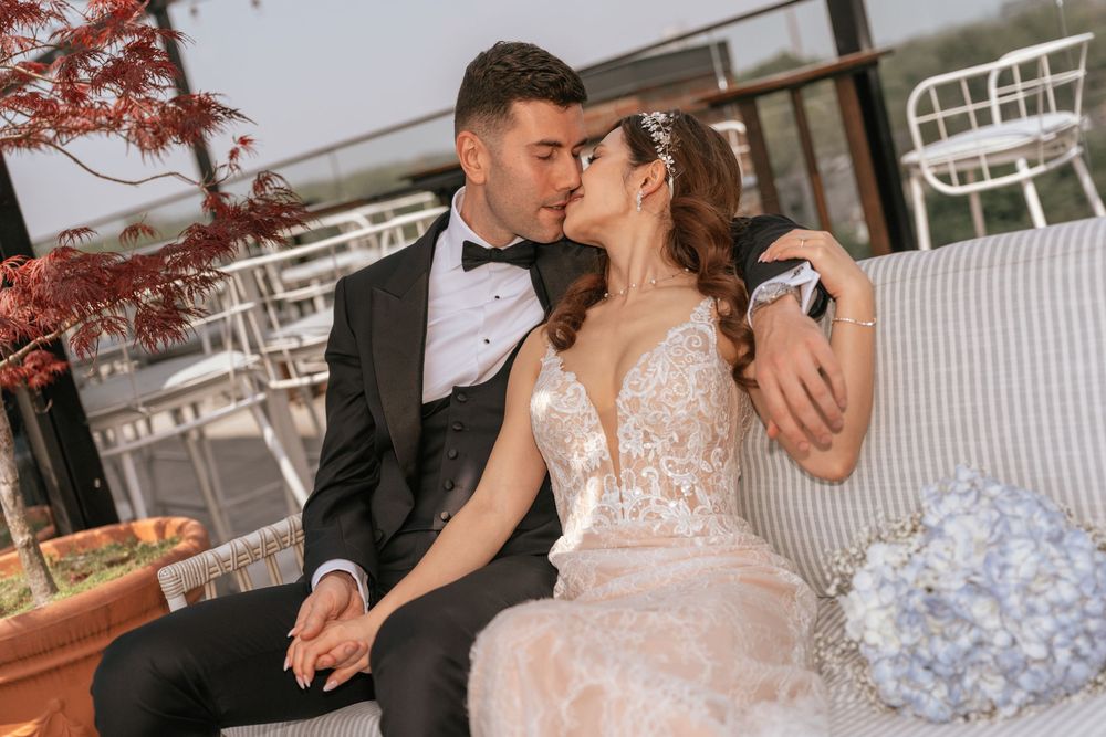 Bride and groom sharing a romantic kiss while seated on a sofa, bride in lace gown, groom in black tuxedo.