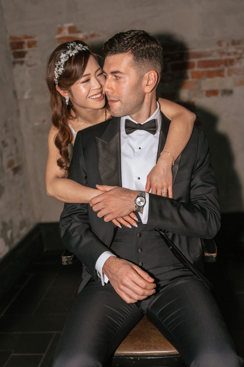 Bride in white dress embraces groom in black tuxedo with bow tie, posing together in romantic wedding portrait.