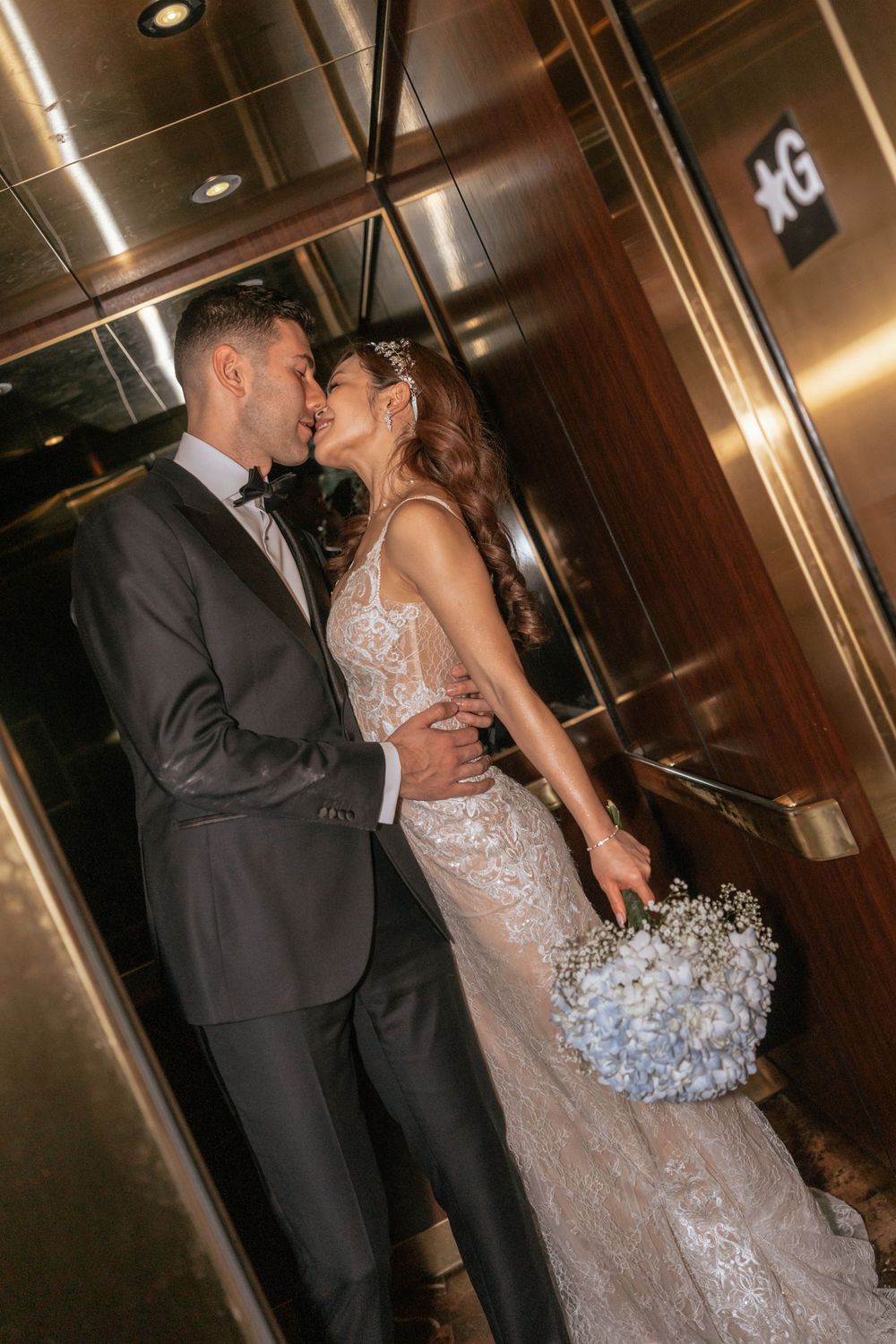 Bride and groom share a romantic kiss in an elegant elevator, bride holding a blue hydrangea bouquet.