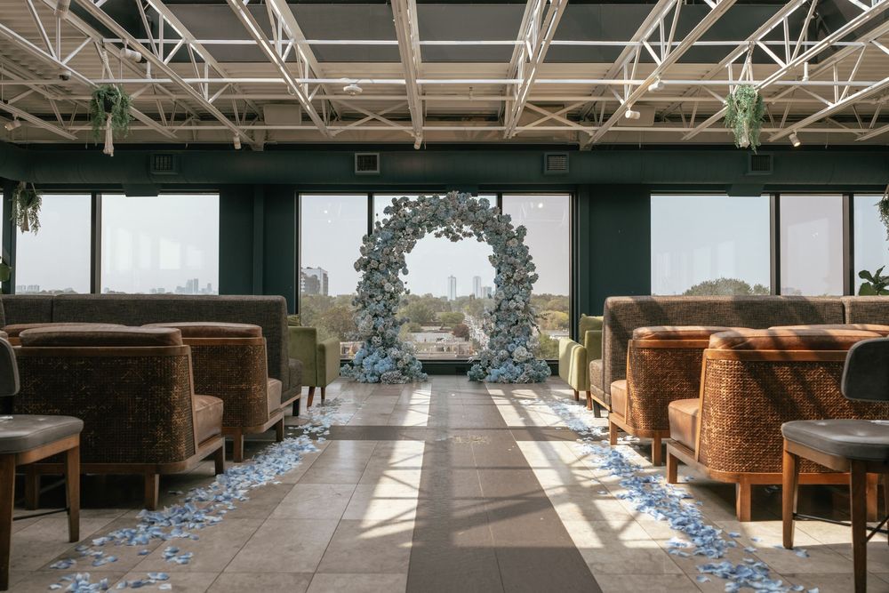 Elegant indoor wedding ceremony with floral arch, petal-lined aisle, and brown lounge chairs in a modern venue.