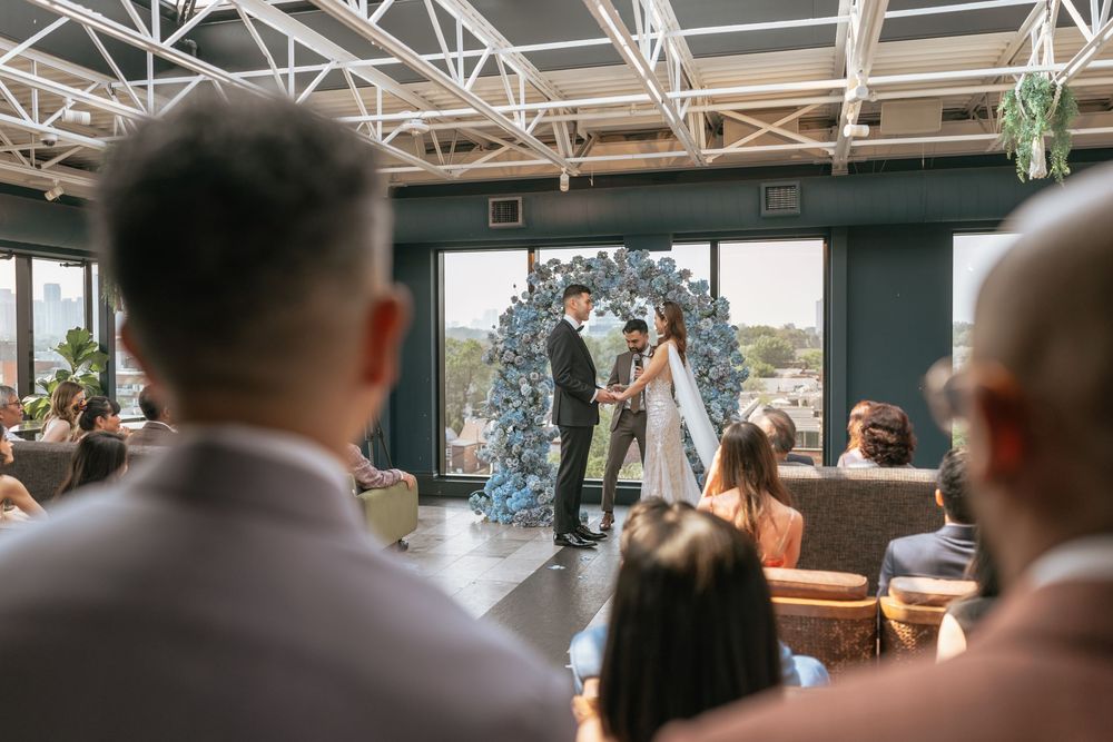 Couple exchanges vows at elegant indoor wedding ceremony with blue floral arch and large windows.