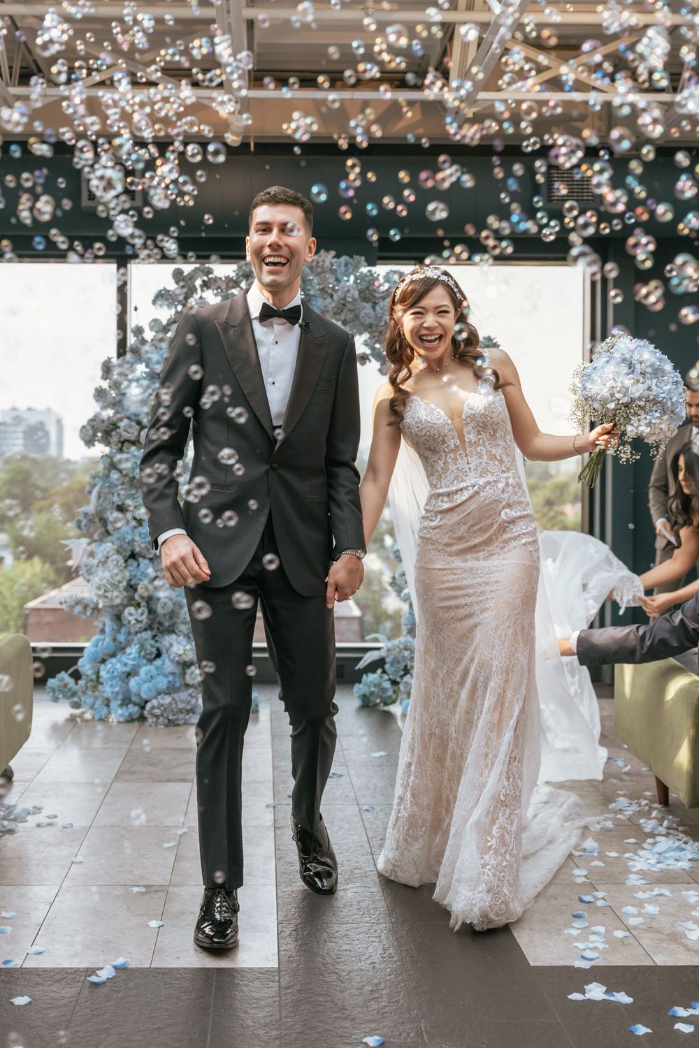 Joyful newlyweds walk through bubbles after wedding ceremony, bride in lace gown holding bouquet, groom in black tuxedo.