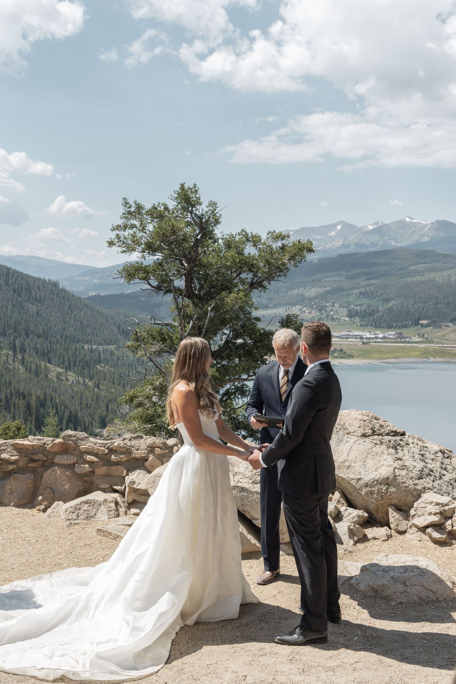 Outdoor mountain wedding ceremony with bride and groom holding hands overlooking a scenic lake and Rocky Mountain landscape.