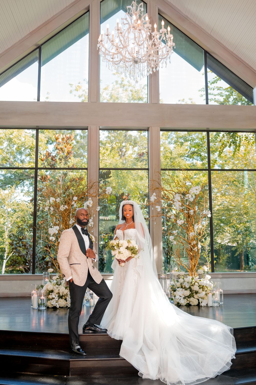 Wedding couple shares romantic moments in a sunlit venue with floor-to-ceiling windows and elegant chandeliers.