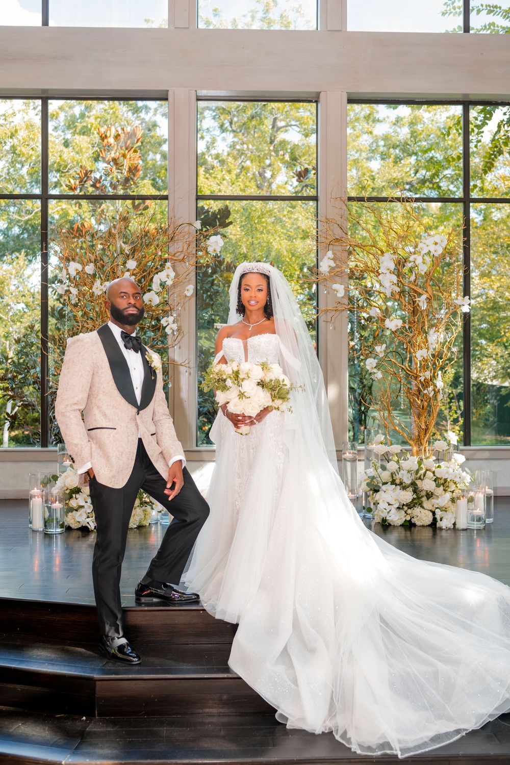Elegant wedding couple poses in front of large windows with fall foliage backdrop in modern venue with floral decorations.