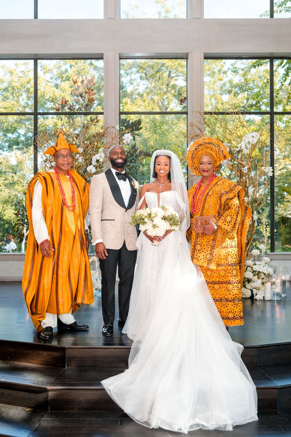 A wedding party in vibrant orange traditional attire poses with the bride in a white gown against a bright windowed backdrop.