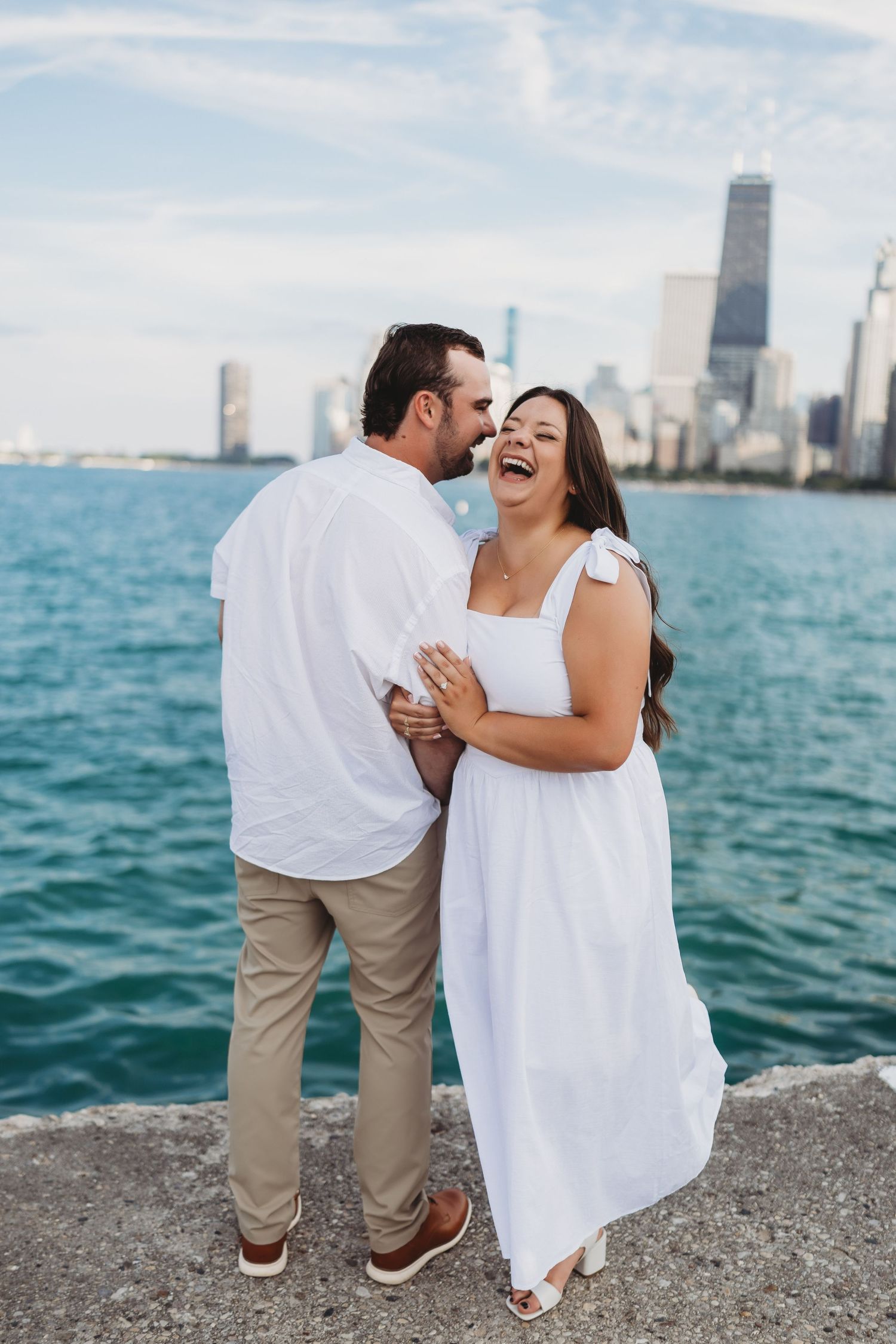 Black and white photo of a couple embracing on a beach, woman's hair blowing in the wind with city skyline behind.