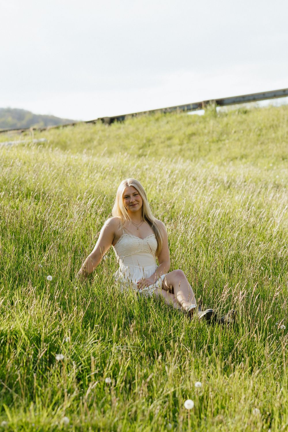 Blonde woman in white dress sitting in green grass field smiling at camera on cloudy day.