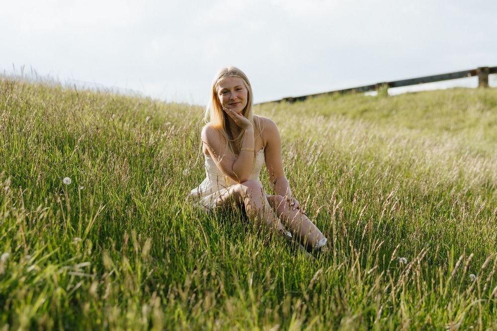 Young blonde woman in a beige dress sitting on a lush green grassy hillside, smiling outdoors on a cloudy day.