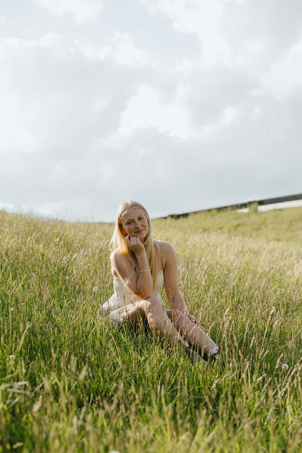 Smiling blonde woman in white dress sitting in green grass field under cloudy sky.