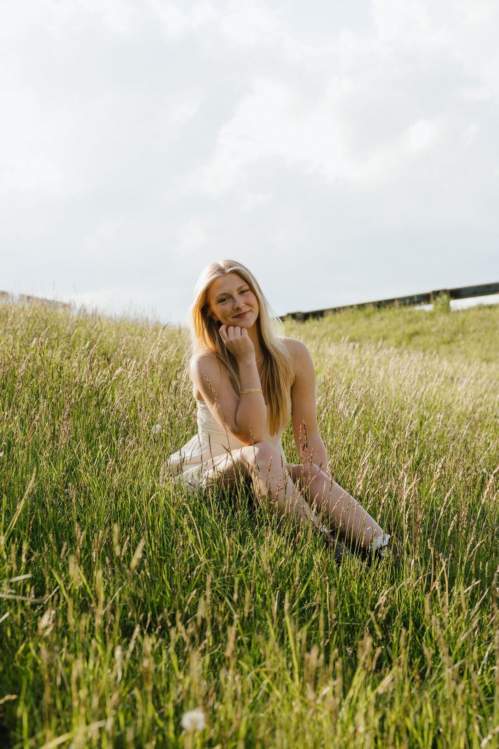 Young blonde woman sitting in lush green meadow wearing white dress on overcast day.