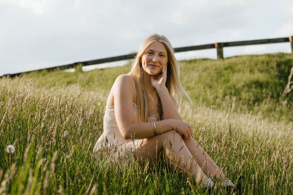 Young blonde woman sitting in lush green grass field, smiling peacefully in a light pink dress on a cloudy day.