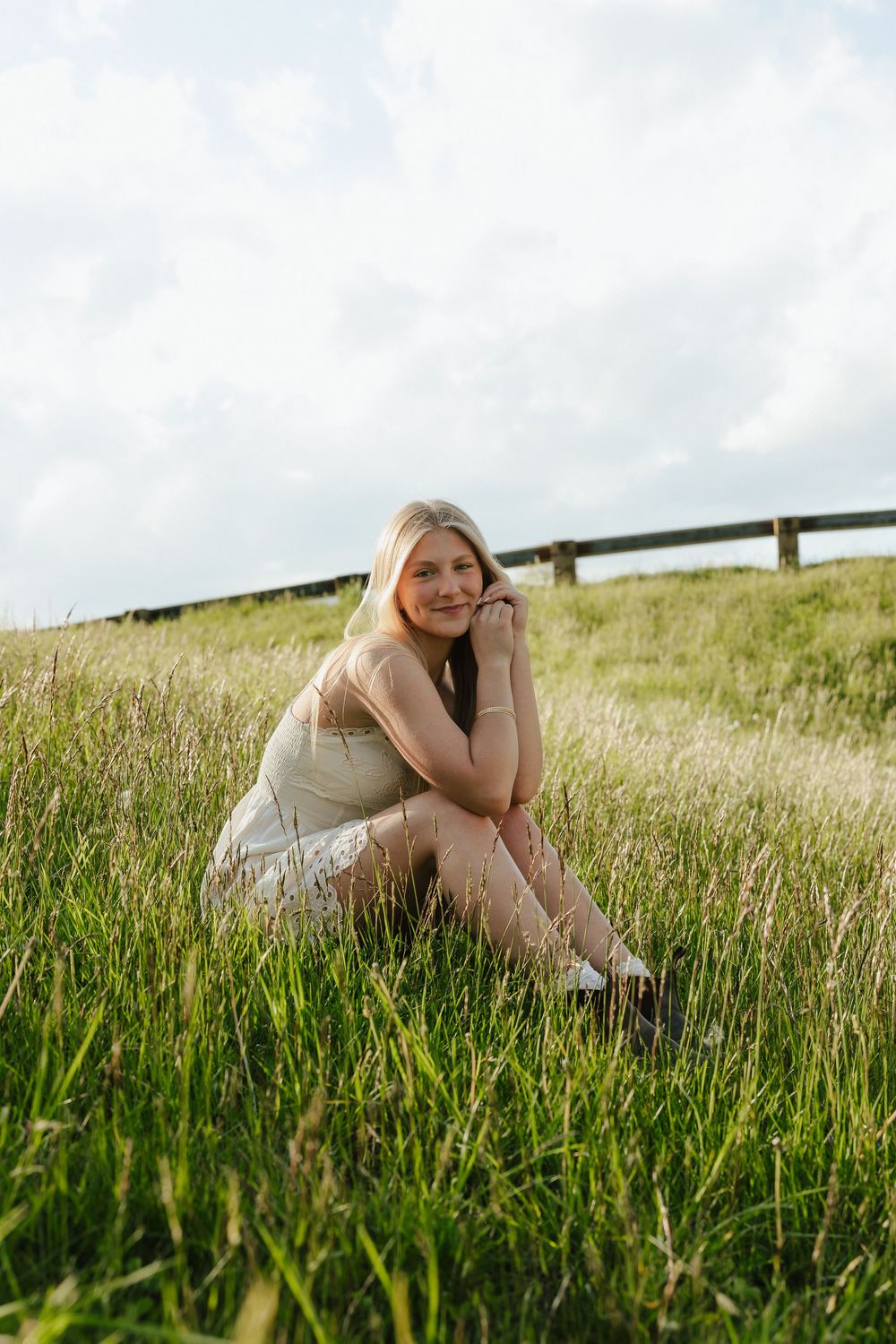 Young blonde woman in white dress sitting relaxed in lush green meadow with wooden fence.