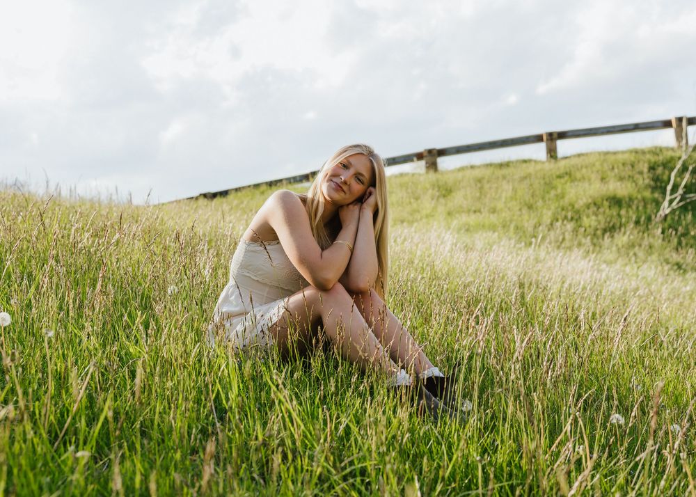 Young blonde woman in a beige dress sitting in tall green grass field with a wooden fence and cloudy sky.