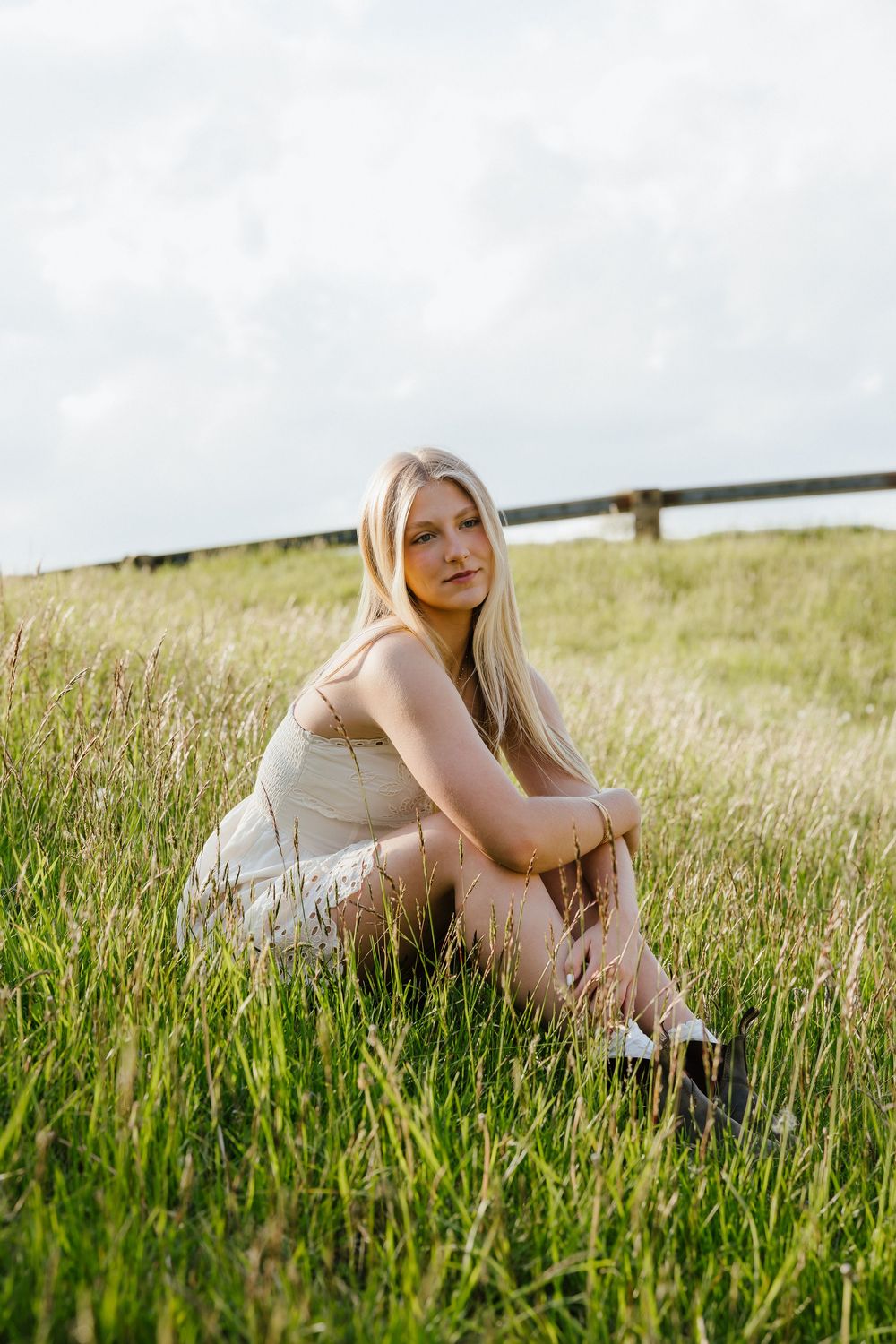 Blonde teen girl in white dress sitting in green grass field for senior portrait photography.