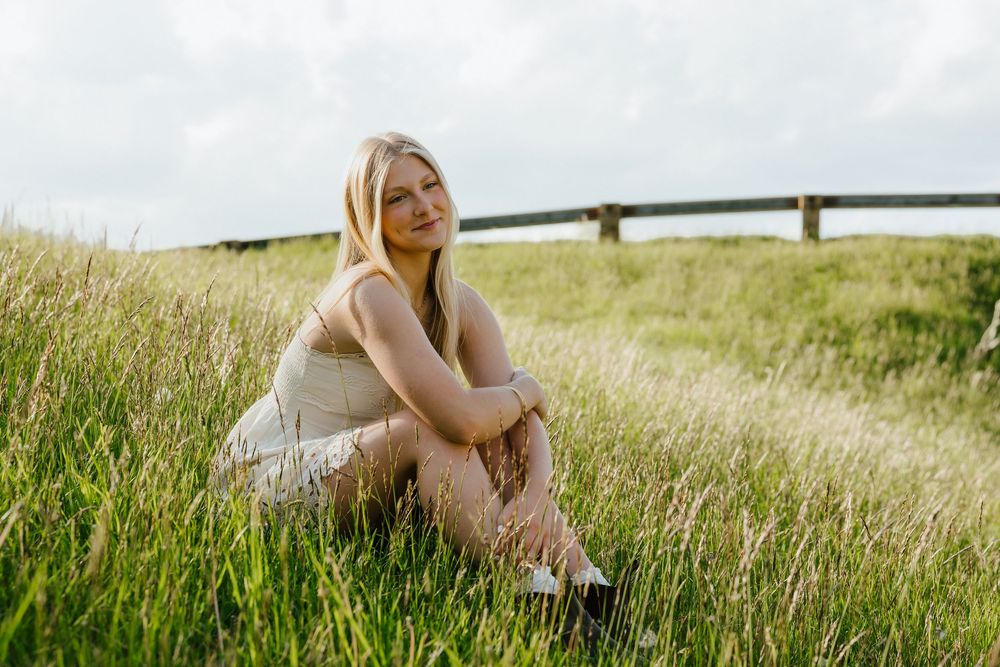 Young blonde woman sitting in tall green grass field wearing a white dress, smiling softly on a cloudy day.