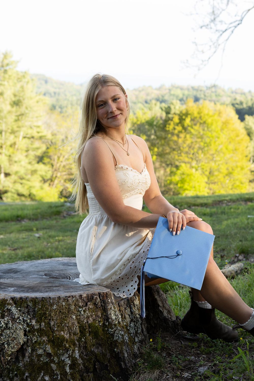 Blonde graduate in white dress sitting on tree stump holding light blue graduation cap outdoors.