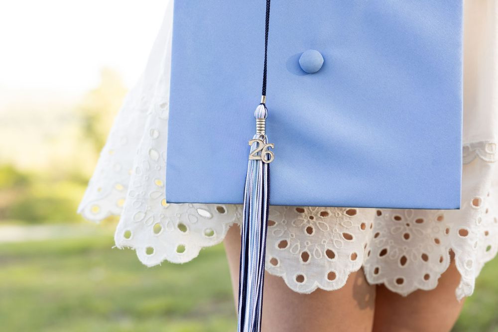 Close-up of a light blue graduation cap with tassel and '26 charm, held by a graduate in a white lace dress.