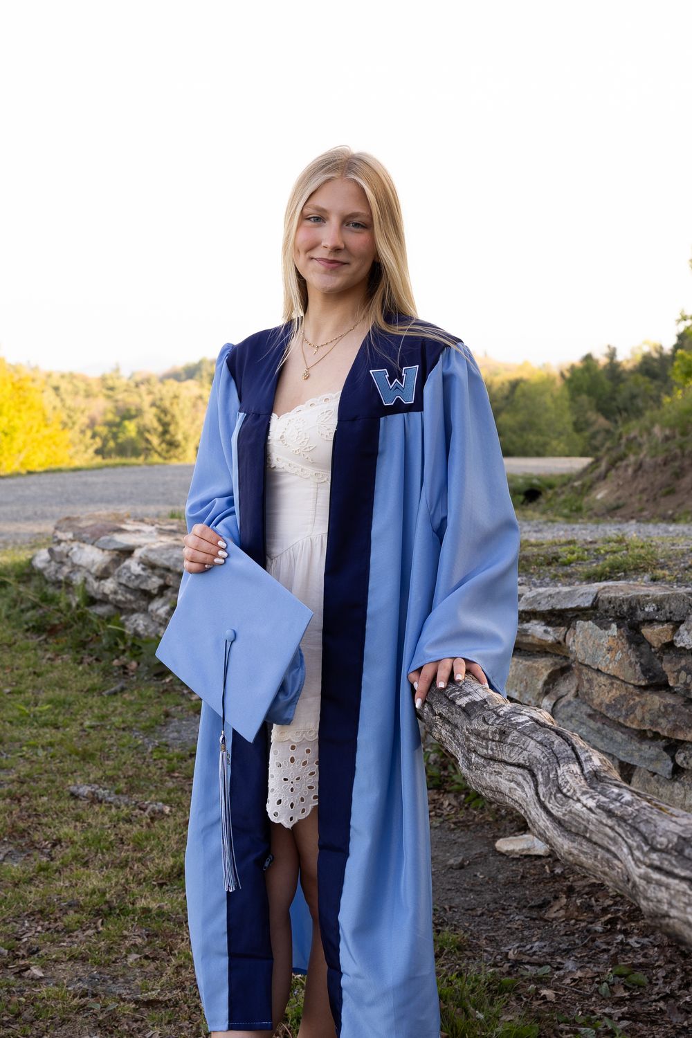Blonde graduate in light blue cap and gown standing outdoors near stone wall at sunset.