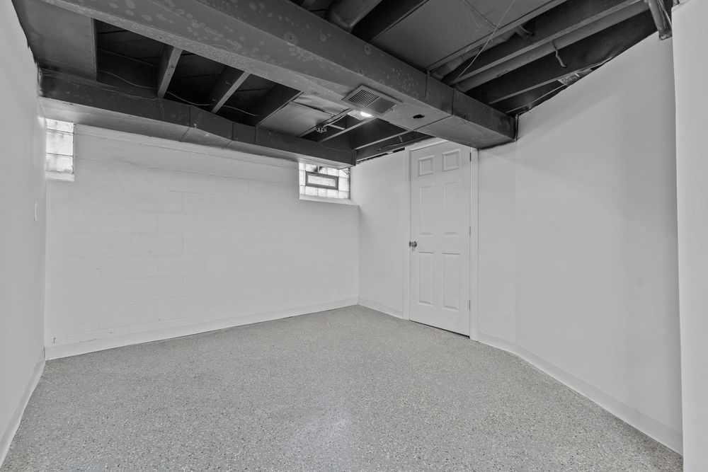 Unfinished basement with white painted walls, exposed black ceiling joists, epoxy speckled floors, and a white door. Detroit MI Real Estate